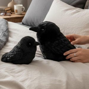May include: Two plush crow toys with black, textured fabric that mimics feathers. Both birds have dark, round eyes and pointed beaks. The larger crow is positioned on the right, while the smaller crow is situated to its left on a light-coloured fabric surface.