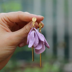 May include: A pair of drop earrings featuring gold-tone metal posts with a spiral design, attached to clusters of soft lavender-coloured petal-shaped elements and slender, straight gold-tone metal bars hanging at the base of each earring.