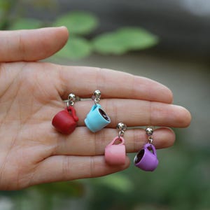 Op de afbeelding: Vier miniatuur oorknopjes in de vorm van kleine koffiemokken in rood, lichtblauw, lichtroze en paars. Elke mok is voorzien van een zilverkleurige metalen stift en een klein oortje, hangend aan een ronde zilverkleurige kogelvormige sluiting.