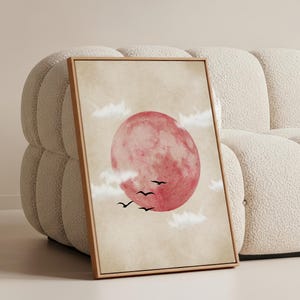 Puede incluir: Una impresión artística rectangular enmarcada que presenta una gran luna circular de color rojo rosado y texturizada, centrada sobre un fondo beige apagado. Varios pájaros pequeños en silueta negra vuelan sobre la parte inferior de la luna, rodeados de nubes blancas suaves y tenues.
