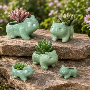 May include: A collection of five light green ceramic planters shaped like four-legged creatures. Three of the planters contain small green or pink succulents, while two are empty. The planters feature smooth, glossy surfaces and rounded ears.