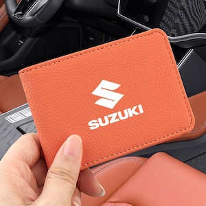 Peut inclure: Un portefeuille rectangulaire orange à la surface texturée, arborant le logo et le texte Suzuki en blanc au centre. Le portefeuille présente des coutures visibles sur les bords et est tenu de manière à bien montrer la face avant.