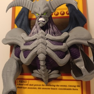 Può includere: Una figura stampata in 3D di un mostro cornuto con armatura grigia, busto viola e artigli neri, montata davanti a una carta collezionabile gialla con il testo FIEND, A fiend with dark powers for confusing the enemy, ATK/2500 e DEF/1200.