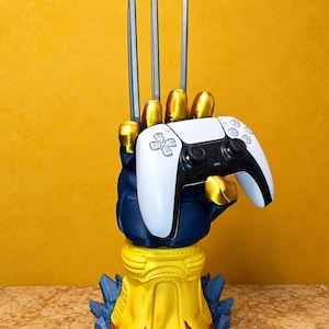 Peut inclure: Un support de manette en forme de main bleue et jaune avec trois longues griffes métalliques argentées pointant vers le haut. La main tient une manette de jeu vidéo sans fil blanche et noire avec des boutons et des joysticks sur sa face avant.