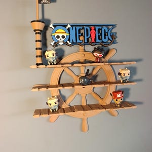 Könnte beinhalten: Ein beigefarbenes Wandregal in Form eines Schiffssteuerrads mit drei horizontalen Ablageflächen. Oben befindet sich ein One Piece-Logo mit Totenkopf, ein Mast mit schwarzer Flagge und fünf kleine Charakterfiguren auf den Regalböden.