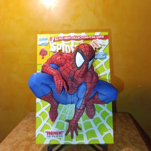 Pode incluir: Uma figura tridimensional do Spider-Man em pose agachada, montada sobre uma capa de banda desenhada. A figura apresenta um fato vermelho e azul com padrões de teia pretos e olhos brancos grandes. A capa exibe o texto Spider-Man, Torment e 1st All-New Collectors Item Issue.
