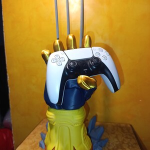 May include: A controller stand shaped like a blue and yellow gloved hand with gold fingertips and three silver claws extending upward. The hand is holding a white and black wireless game controller. The base features blue jagged rock-like accents.