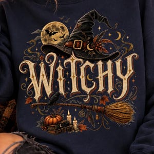 May include: A dark navy blue crewneck sweatshirt featuring a graphic print with the word WITCHY in large decorative lettering. The design includes a black witch hat, a broomstick, a full moon with bats, a pumpkin, candles, and books.