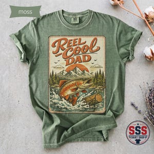 May include: A moss green short-sleeve t-shirt featuring a graphic print of a jumping fish, a log, mountains, and trees. The text "REEL COOL DAD" is printed in a stylized brown font above the landscape illustration, which includes a sunset.