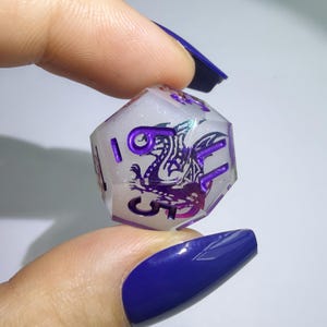 May include: A translucent, multi-faceted polyhedral die with a milky white, glitter-infused base. The die features a detailed purple dragon graphic and deep purple engraved numbers, including a prominent number six, across its various faces.