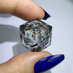 May include: A twenty-sided die made of clear resin containing iridescent glitter and small black inclusions. The numbers are engraved in a metallic copper colour, with the number eleven prominently displayed in the centre of the front face.
