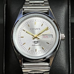 May include: A silver-toned Citizen automatic wristwatch featuring a metallic silver dial with gold-colored hour markers and hands. The watch includes a day-date window at the three o'clock position, a logo with the number 7, and a stainless steel link bracelet.