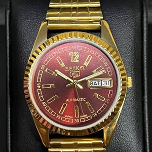 May include: A gold-tone Seiko 5 automatic wristwatch featuring a deep red sunburst dial with gold-tone indices and hands. The watch includes a day-date window at the three o'clock position, a fluted bezel, and a matching gold-tone metal link bracelet.