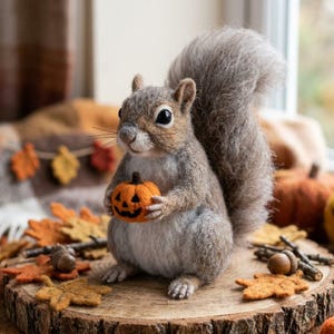 May include: A needle-felted grey squirrel figurine with a bushy tail, holding a small orange pumpkin with a carved jack-o'-lantern face. The squirrel has dark eyes, a light-coloured chest, and is positioned on a round wooden slice base.