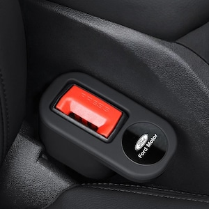 May include: A black car seat belt buckle extender featuring a bright red release button with the word PRESS embossed on it. The unit includes a circular black emblem with the white Ford logo and the text Ford Motor printed in a clear font.