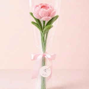 Könnte beinhalten: Eine einzelne gehäkelte rosa Pfingstrose mit grünen Blättern und Stiel, verpackt in durchsichtiger Zellophanfolie. Der Strauß ist mit einem hellrosa Satinband gebunden und hat ein kleines rundes weißes Etikett mit Blumenskizze und dem Text For you.