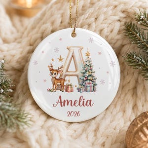 Peut inclure: Une décoration ronde en céramique blanche ornée d'une grande lettre A dorée, d'un petit cerf brun, d'un sapin de Noël décoré et de boîtes cadeaux colorées. Le prénom Amelia est écrit en rose sous la lettre, suivi de l'année 2026.