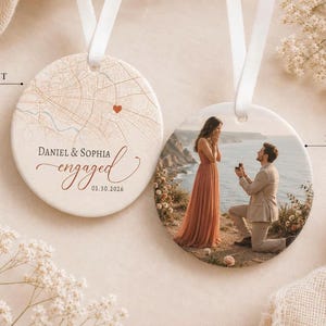 May include: Two round ceramic ornaments with white ribbons. The front features a tan map with a red heart, the text "DANIEL & SOPHIA engaged" and "01.30.2026". The back displays a photograph of a person proposing to another on a cliffside.