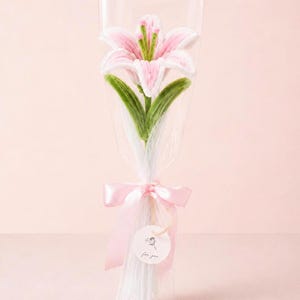 Puede incluir: Una flor de lirio de ganchillo en tonos rosa y blanco con hojas verdes, envuelta en una película de plástico transparente. El ramo está atado con una cinta de satén rosa claro y tiene una pequeña etiqueta redonda blanca con un dibujo floral.