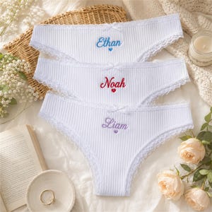May include: Three white ribbed cotton panties featuring delicate lace trim along the edges and a small bow at the center waist. Each pair is embroidered with a name in cursive script: Ethan in blue, Noah in red, and Liam in purple, each with a heart.