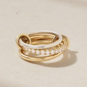 May include: A multi-band ring featuring a combination of polished gold-tone and silver-tone metal bands. One of the central bands is encrusted with a row of small, clear, round-cut stones that catch the light, creating a layered and interconnected design.