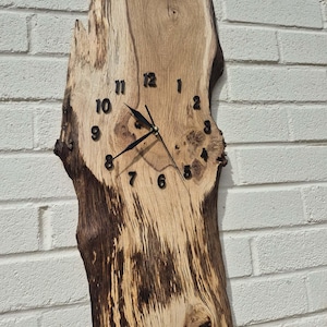 Op de afbeelding: Een rustieke wandklok gemaakt van een natuurlijke houten plank. De klok heeft zwarte cijfers en wijzers, tegen de natuurlijke nerf en kleurvariaties van het hout. De onregelmatige vorm van de klok draagt bij aan de unieke, organische uitstraling.