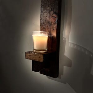 Peut inclure: Une applique murale en bois brun foncé composée d'un panneau arrière rectangulaire vertical au grain de bois visible et d'une petite étagère horizontale. Un bougeoir en verre transparent contenant une bougie blanche allumée est posé dessus.
