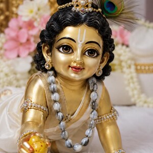 Könnte beinhalten: Eine goldene Figur einer krabbelnden Kindgottheit mit dunklem, gewelltem Haar, geschmückt mit einem perlenbesetzten Kopfschmuck und einer Pfauenfeder. Die Figur trägt ein weißes Gewand, eine marmorierte Perlenkette und Armbänder aus Perlen.