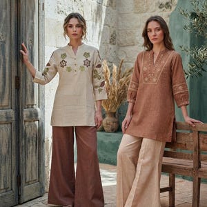 May include: Two outfits consisting of long-sleeved tunics and wide-leg trousers. One tunic is beige with floral embroidery and brown trousers. The other is a brown tunic with white embroidery and beige trousers. Both feature V-necklines.