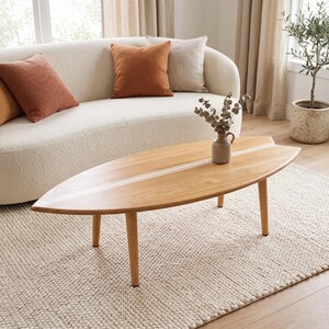 Op de afbeelding: Een surfboardvormige salontafel gemaakt van lichtgekleurd hout, voorzien van een centrale witte inlegstrip over de gehele lengte. De tafel rust op vier taps toelopende houten poten en bevat een kleine keramische vaas met groen blad.