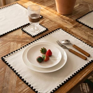 Pode incluir: Um conjunto de jogos americanos retangulares em tom branco sujo com uma borda decorativa preta recortada. O conjunto inclui porta-copos quadrados a condizer, pratos de cerâmica branca, um copo estriado e talheres de aço com cabos de madeira.