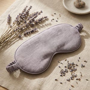 May include: A lavender-colored sleep mask made of woven fabric with a matching elastic strap. The mask features a smooth, contoured shape designed to cover the eyes, with visible fabric texture and a simple, clean edge finish.