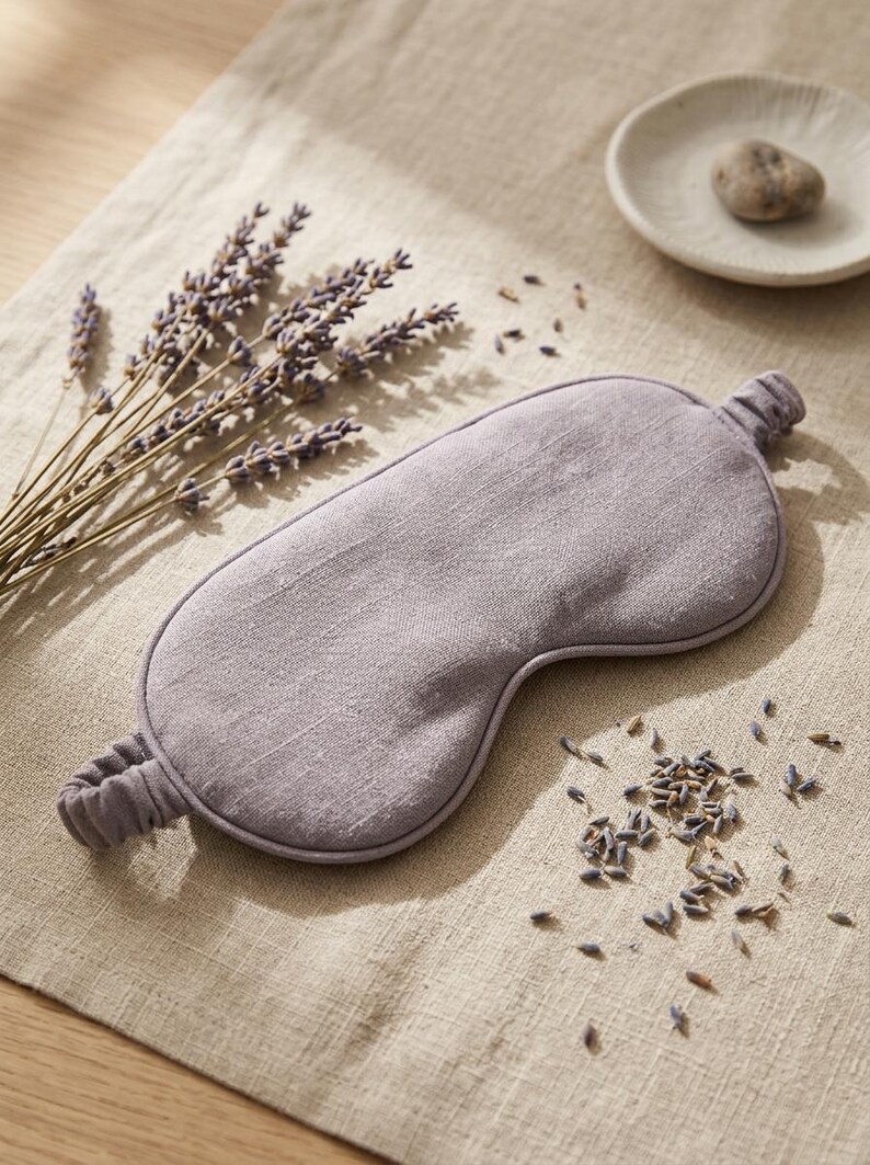 May include: A lavender-colored sleep mask made of woven fabric with a matching elastic strap. The mask features a smooth, contoured shape designed to cover the eyes, with visible fabric texture and a simple, clean edge finish.