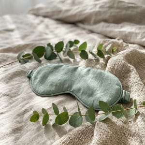 May include: A sage green sleep mask with a textured fabric finish and a gathered elastic strap. The mask features a contoured shape designed to cover the eyes, showcasing a visible weave pattern in the material and a soft, muted color tone.