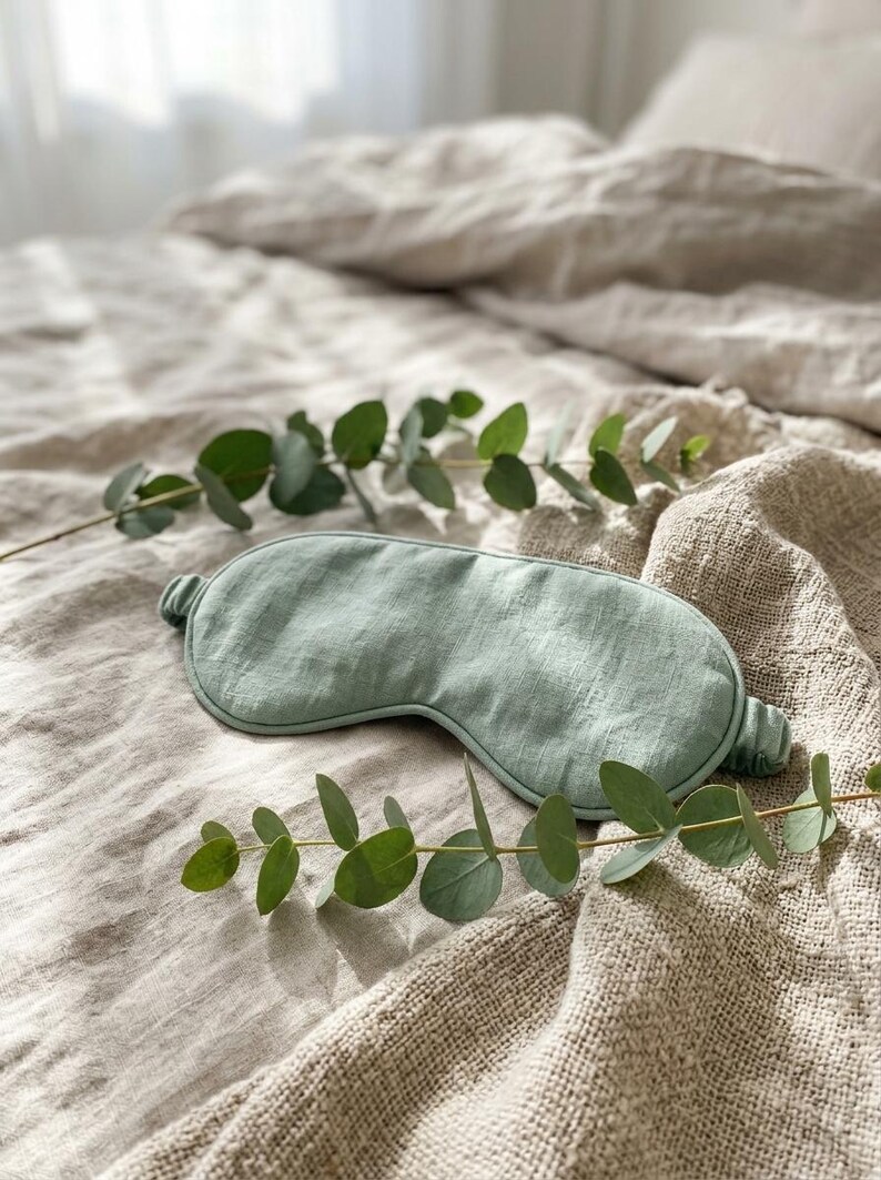 May include: A sage green sleep mask with a textured fabric finish and a gathered elastic strap. The mask features a contoured shape designed to cover the eyes, showcasing a visible weave pattern in the material and a soft, muted color tone.