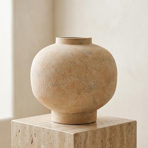 Puede incluir: Un jarrón esférico redondo con un cuerpo ancho y bulboso y un cuello corto y estrecho. La superficie tiene un acabado mate y texturizado en tonos beige claro, arena y naranja suave, que recuerda a la piedra natural con motas sutiles.