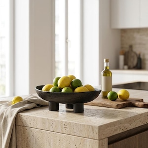 May include: A dark, textured, oval-shaped bowl with three short, cylindrical legs, filled with a collection of bright yellow lemons and green limes. The bowl is positioned on a light-colored stone surface next to a glass bottle of oil.