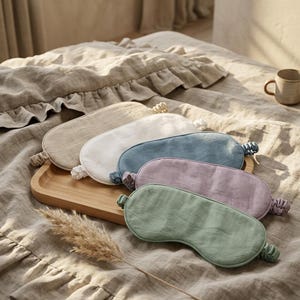 May include: A collection of five oval-shaped sleep masks made of linen fabric, each featuring an elasticated strap. The masks are presented in a variety of solid colors including beige, white, blue, lavender, and sage green, arranged on a wooden tray.