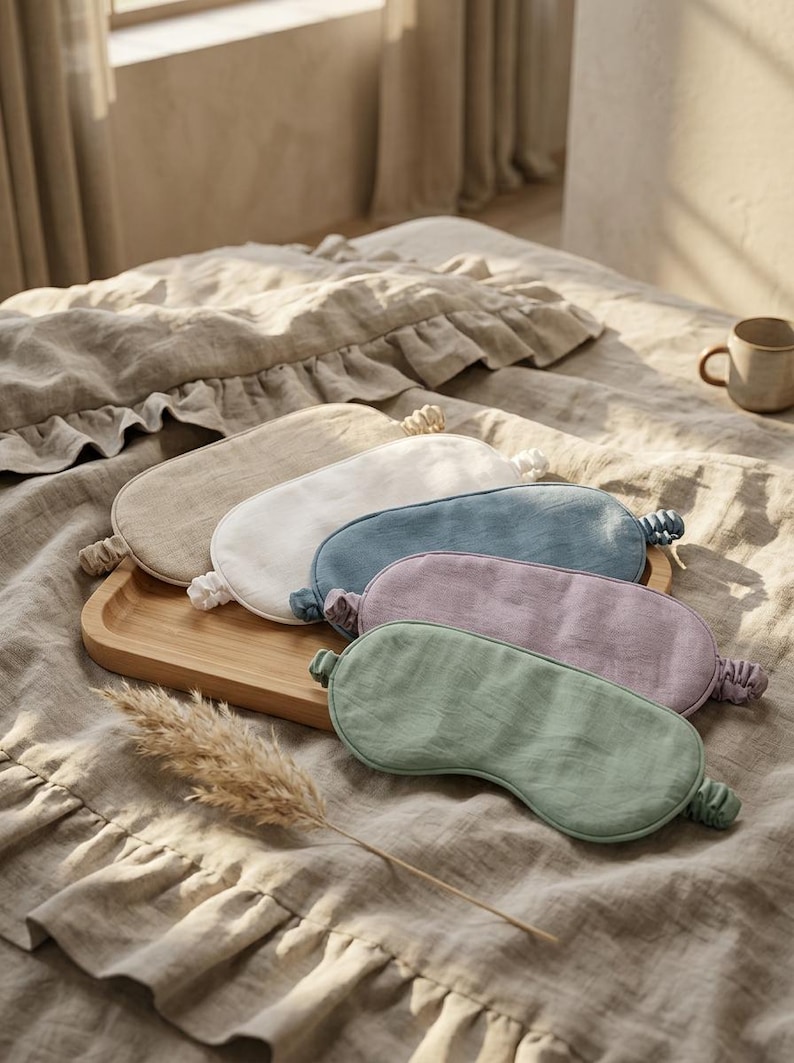 May include: A collection of five oval-shaped sleep masks made of linen fabric, each featuring an elasticated strap. The masks are presented in a variety of solid colors including beige, white, blue, lavender, and sage green, arranged on a wooden tray.