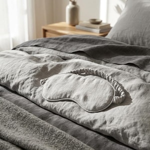 May include: A light gray sleep mask with a textured fabric finish and a matching gathered elastic strap. The mask features a contoured shape designed to cover the eyes, resting on a gray linen bedspread with a visible woven fabric texture.
