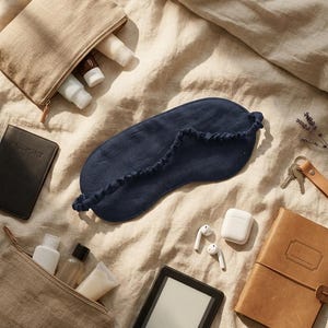 May include: A dark blue linen sleep mask with a gathered elastic strap, two beige linen toiletry pouches containing small bottles, a black passport holder, a black e-reader, white wireless earbuds in a charging case, and a tan leather notebook.