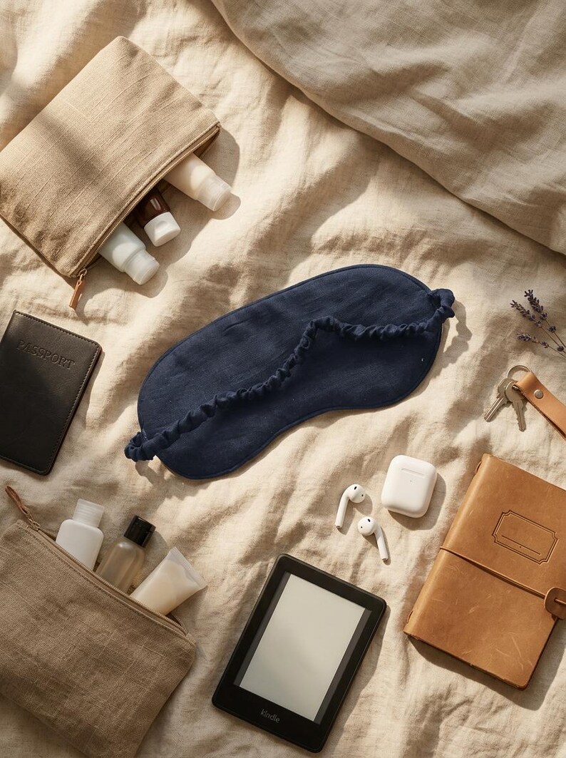May include: A dark blue linen sleep mask with a gathered elastic strap, two beige linen toiletry pouches containing small bottles, a black passport holder, a black e-reader, white wireless earbuds in a charging case, and a tan leather notebook.