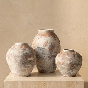 May include: Three ceramic vases with a weathered, textured finish in shades of terracotta, gray, and off-white. The vases feature rounded bodies and narrow openings, arranged in a group with one taller central vase flanked by two smaller ones.