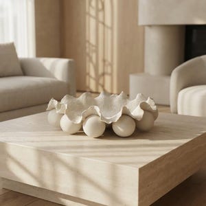 May include: A decorative bowl with a ruffled, shell-like edge in an off-white finish. The bowl is supported by a series of smooth, spherical balls arranged in a circular pattern around the base, creating a raised, sculptural appearance.