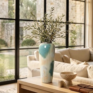 May include: A tall, cylindrical ceramic vase featuring a blended glaze of teal, cream, and soft peach tones. The vase contains a cluster of green olive branches with slender leaves. It has a smooth, glossy finish and a narrow, circular opening.