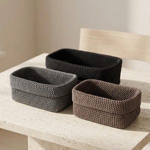 May include: A set of three rectangular storage baskets crafted from thick, crocheted yarn. The baskets are presented in three distinct neutral colors: one in solid black, one in dark charcoal gray, and one in a muted earthy brown tone.