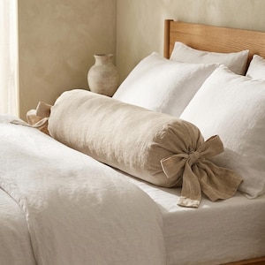 May include: A cylindrical bolster pillow in a light beige color, featuring fabric ties at both ends that are knotted into bows. The pillow is positioned horizontally across a bed covered with white bedding and rests against white pillows.