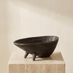 May include: A dark brown, oval-shaped bowl with a rough, textured surface and three short, sturdy legs. The bowl features an uneven, organic rim and a deep interior, presenting a solid, heavy appearance with a matte finish throughout the entire piece.