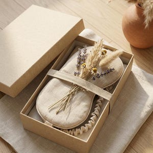 May include: A beige linen sleep mask with an elastic strap, presented in a rectangular cardboard gift box. The mask is decorated with a small bundle of dried lavender sprigs, pampas grass, and tiny yellow chamomile flowers tucked under the strap.