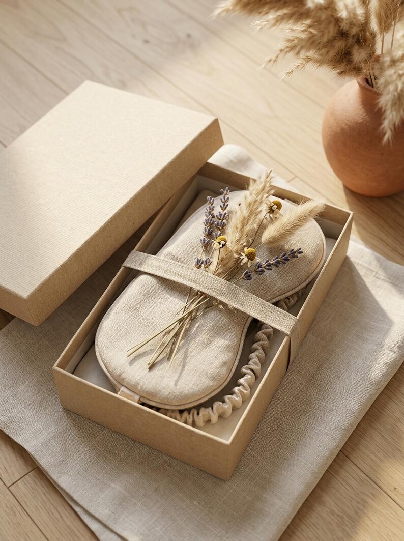 May include: A beige linen sleep mask with an elastic strap, presented in a rectangular cardboard gift box. The mask is decorated with a small bundle of dried lavender sprigs, pampas grass, and tiny yellow chamomile flowers tucked under the strap.